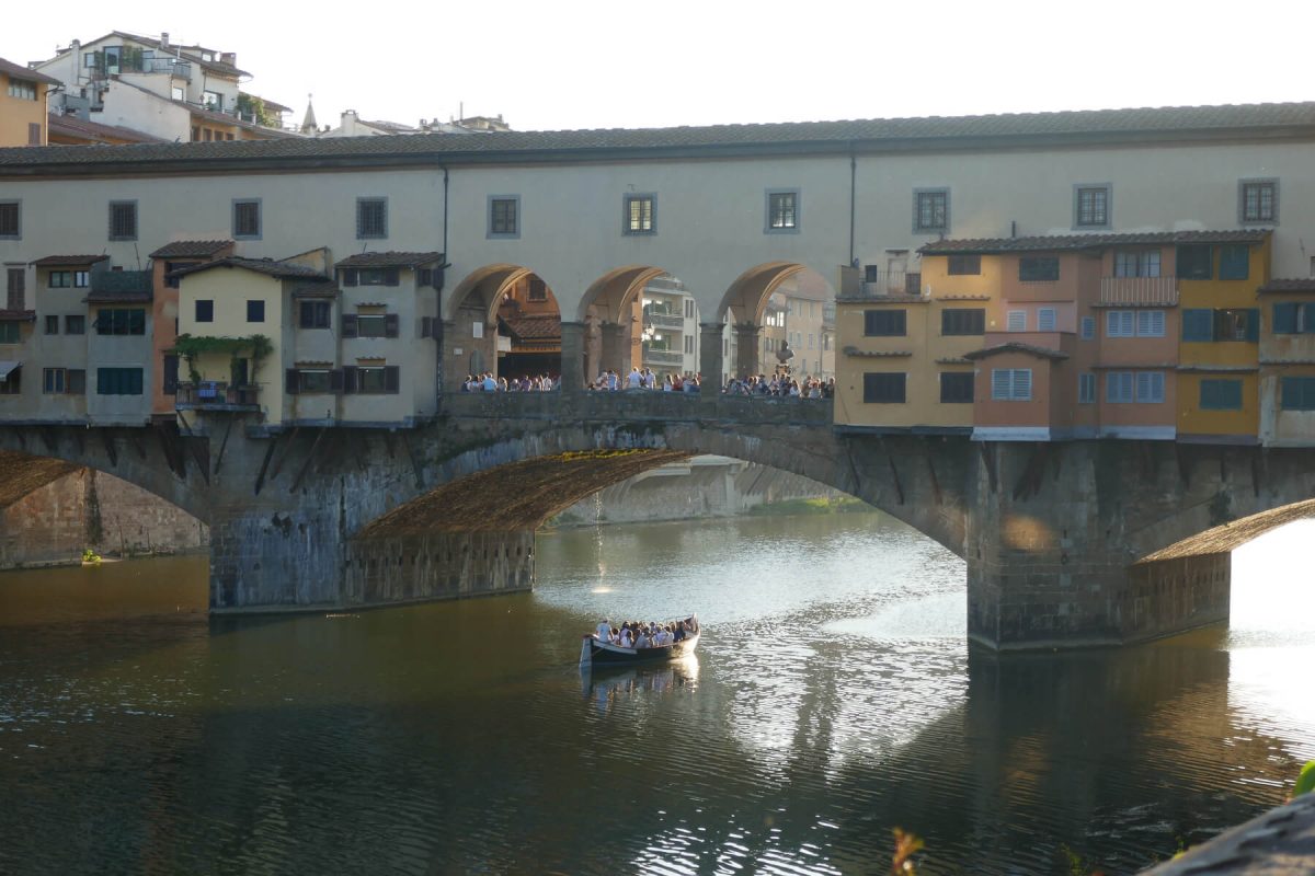 Looking towards an arched bridge with buildings on bridge and people looking over to the river from the arches on the middle of the bridge