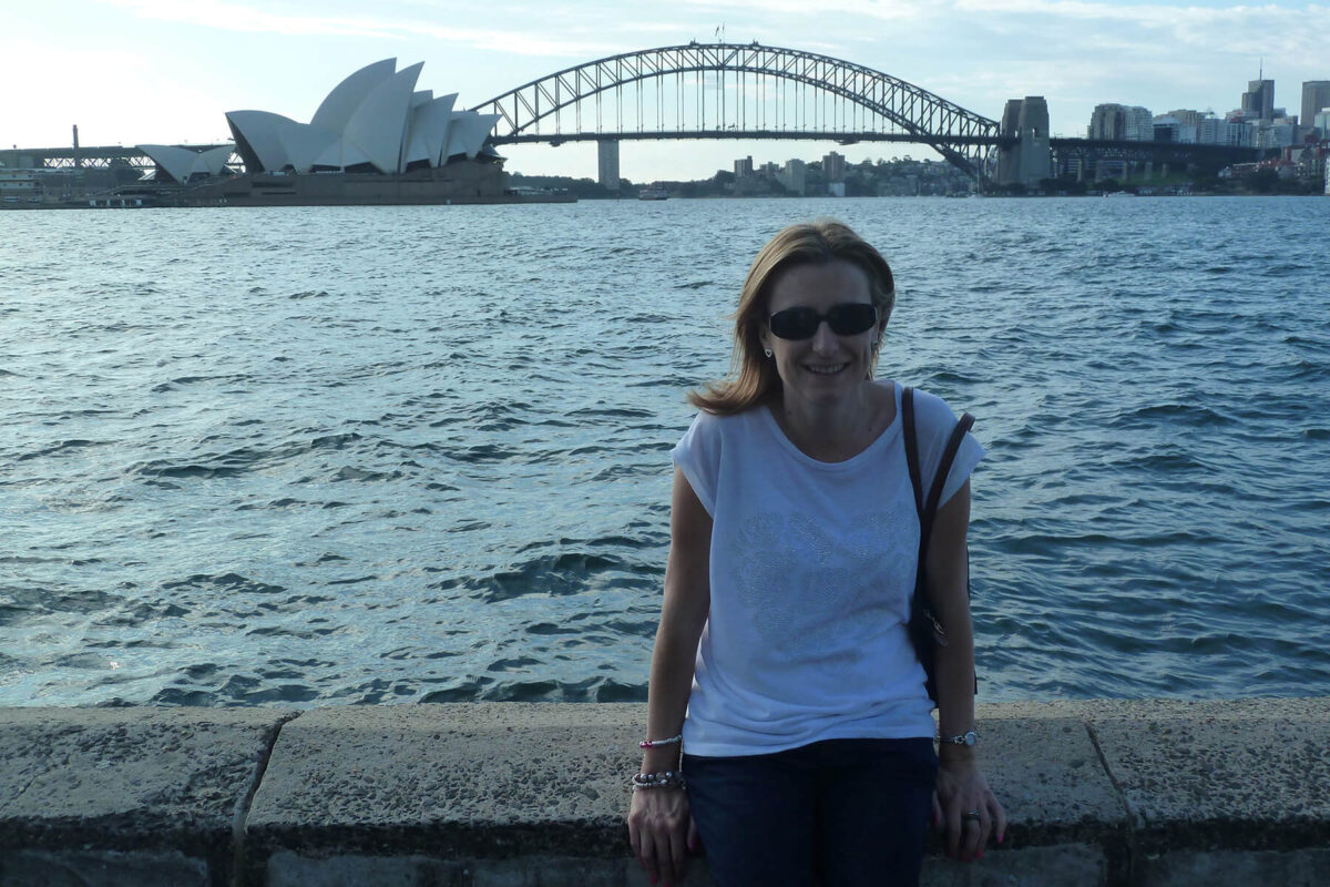 Emma sitting by the water with the Sydney Opera House and Harbour Bridge clearly visible across the harbor in the background.