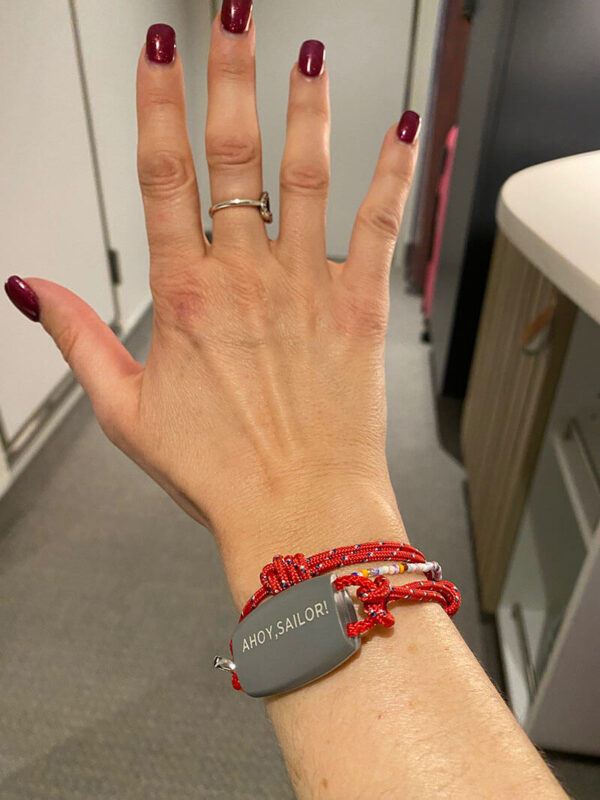 Close-up of a hand wearing a red braided bracelet with a gray tag that reads “AHOY, SAILOR!” in white text. The background shows part of a cruise cabi