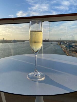 image of a glass of Prosecco on a white table on the deck of a cruise ship overlooking the sea and dock
