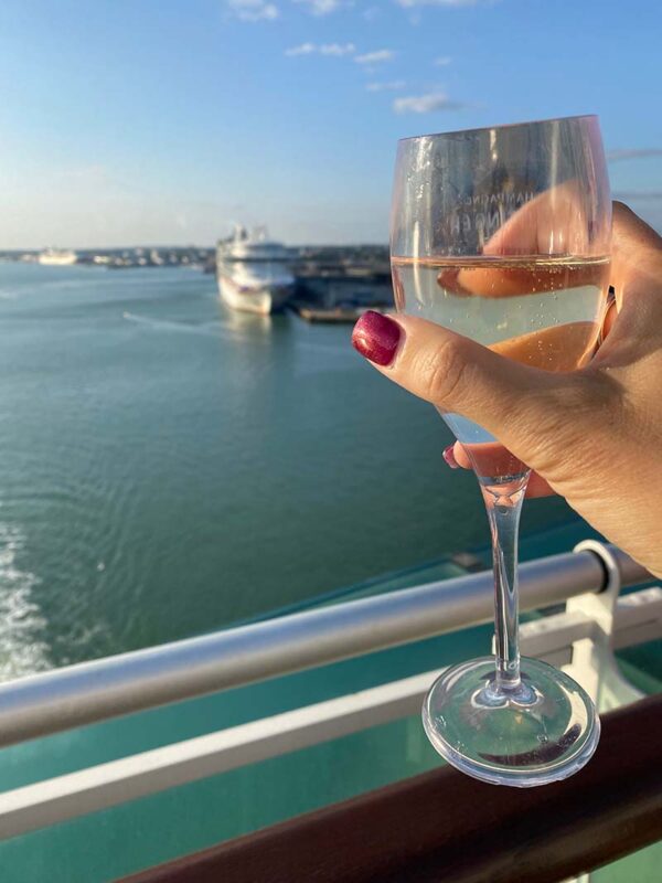 image of a women's hand holding a glass of Prosecco looking out with the sea and a cruise ship docked in the background