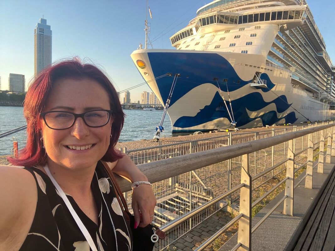Emma posing by the waterfront at sunset with a Princess Cruises ship in the background and a modern city skyline (Rotterdam) rising behind it.