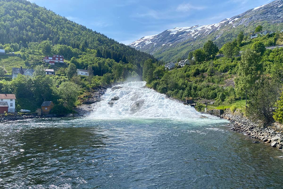 image of a waterfall cascading through the centre of the fjord village of Hellesylt. The waterfall is surrounded by mountain landscape and greenery.