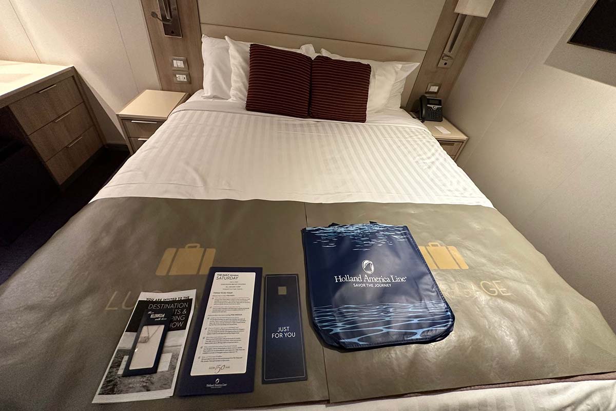 Close-up of a neatly made bed in a Koningsdam stateroom, featuring welcome materials including a blue Holland America Line tote, itinerary pamphlets, and an invitation laid out on a luggage-themed bed runner.