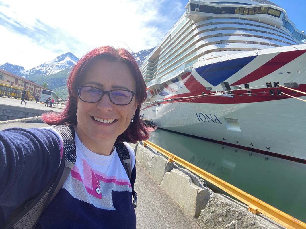 Emma smiling in front of the P&O cruise ship Iona, docked in a scenic fjord port (Olden) with snowy mountains rising in the background.