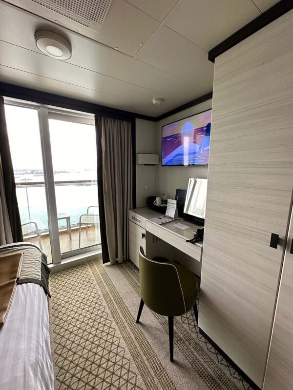 A solo balcony cabin on P&O Cruises Britannia. The bed which is slightly lager than a single but not quite a double, is to the left of the cabin while a desk and chair is to the right. At the end of the cabin is a floor to ceiling sliding door out onto the balcony.