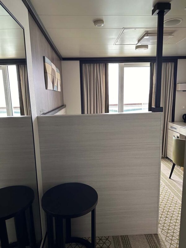 The view as you walk into a solo balcony cabin on P&O Cruises Britannia. In front of you is a small table and the back of the lightly coloured wooden headboard of the bed while to the left is a full sized mirror.