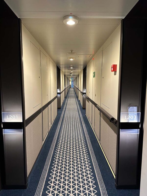 Looking down a long corridor on a cruise ship with cabins either side. The walls are cream with brown wood rails and the carpet is a blue and cream pattern.