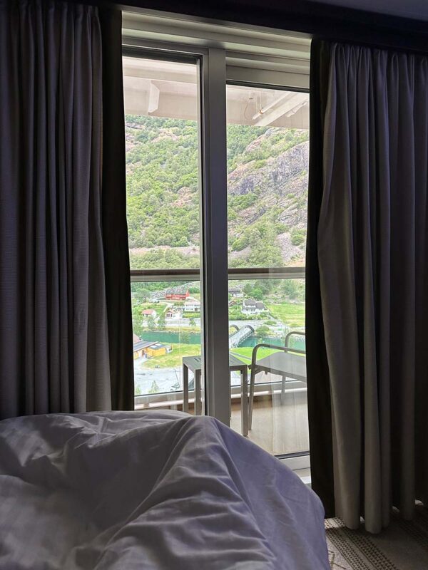 The village of Flam viewed from the bed in a solo balcony cabin on Britannia. A chair and table are on the balcony while in the distance are some buildings in the village and the mountainside.