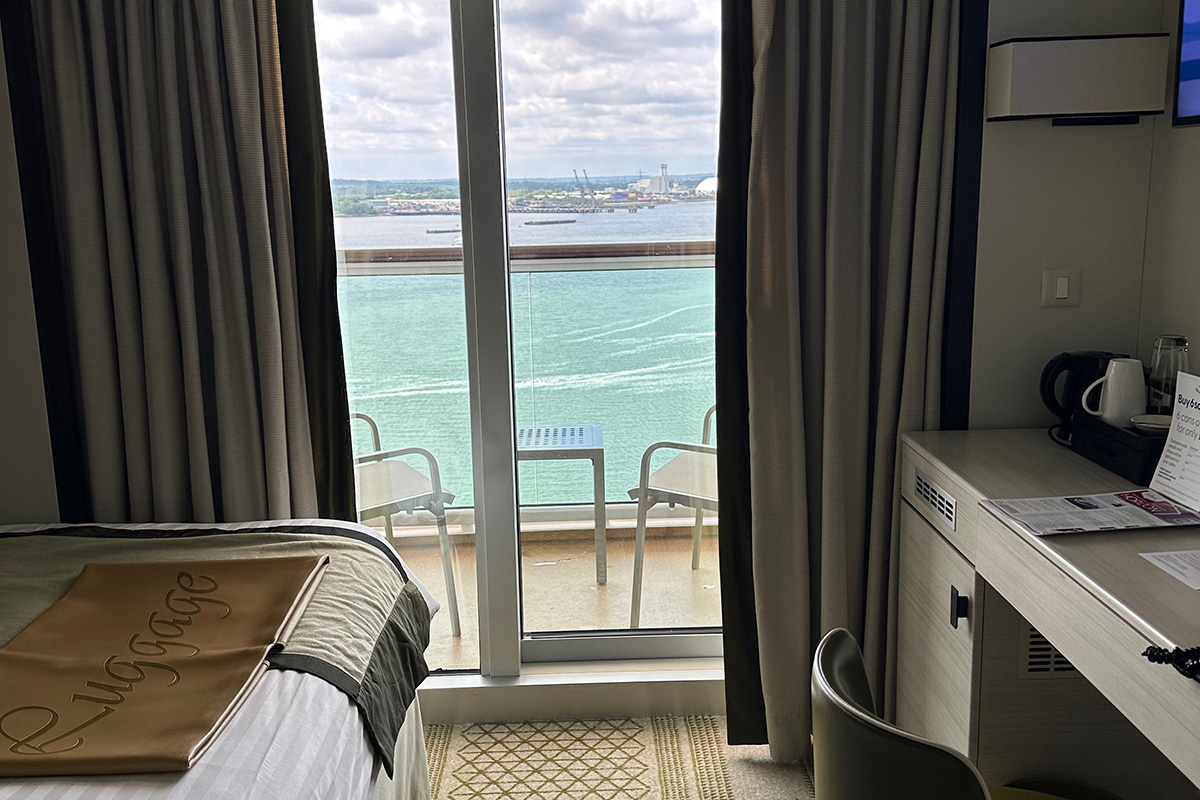 image of part of the solo balcony cabin on P&O Cruises Britannia. In the foreground is part of the single bed and a desk. In the background is the cabin's balcony with a view out to Southampton