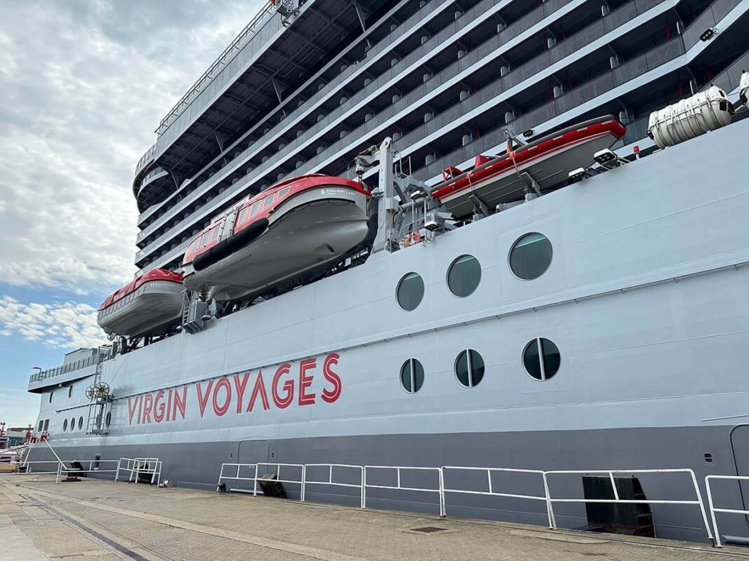 Image of the outside of a Virgin Voyages ship, the hull is grey with the text 'Virgin Voyages' in red alone the side. 