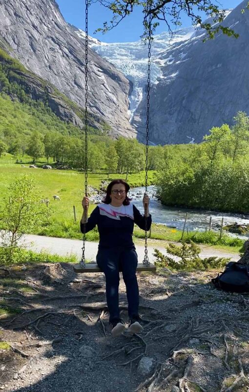 Emma sits smiling on a wooden swing hanging from tree branches, with Briksdal Glacier cascading between steep mountain cliffs in the background. The scene is set in a lush green valley with a small river flowing nearby under a bright blue sky.