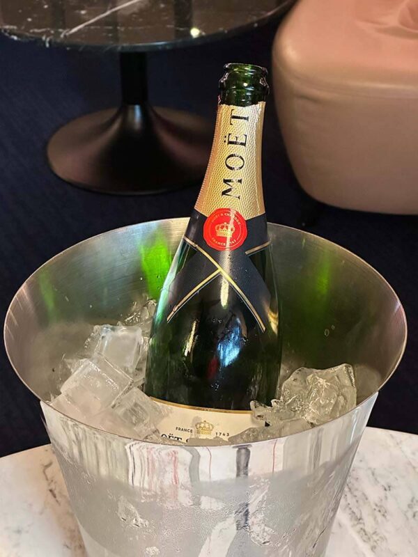A chilled bottle of Moët & Chandon champagne sits in a metal ice bucket filled with large ice cubes on a marble table, with a lounge area in the background.