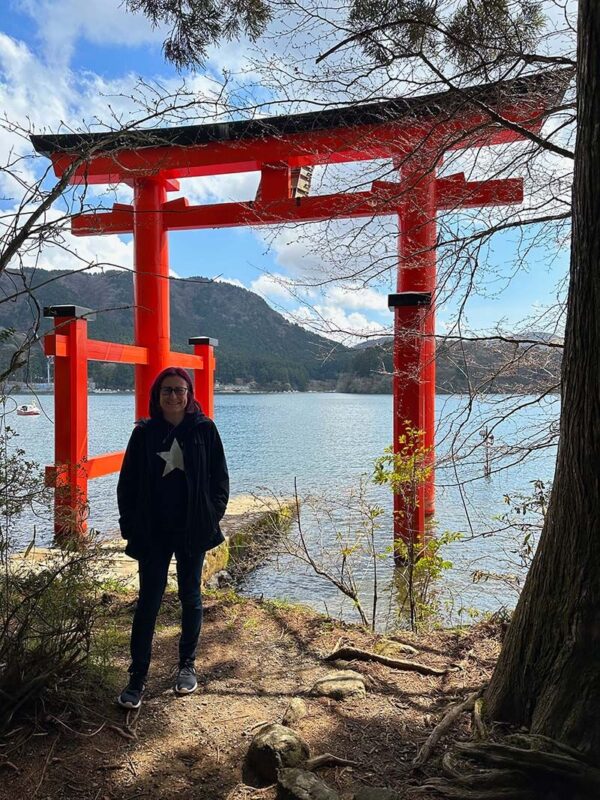 Emma stands in front of a large red torii gate on the edge of a lake, with forested mountains and a bright blue sky in the background. The gate rises from the water, framed by tree branches in the foreground.