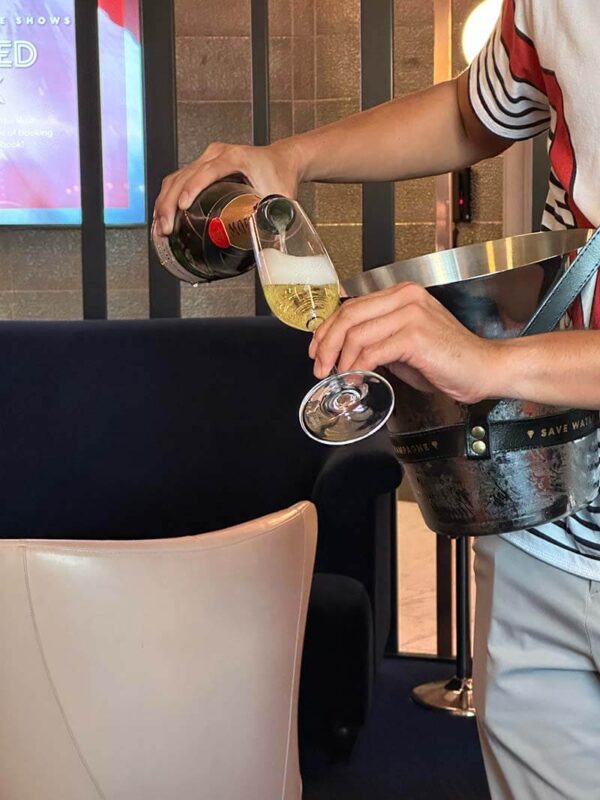 A person wearing a striped shirt pours Moët champagne into a flute while holding an ice bucket strapped over their shoulder in a modern lounge setting.