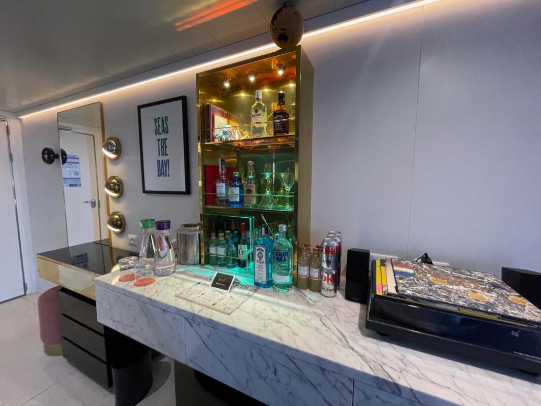 A well-stocked in-room bar with liquor bottles, glassware, and mixers displayed on marble countertops beneath a glowing shelf, with a framed sign reading “SEAS THE DAY!” on the wall.