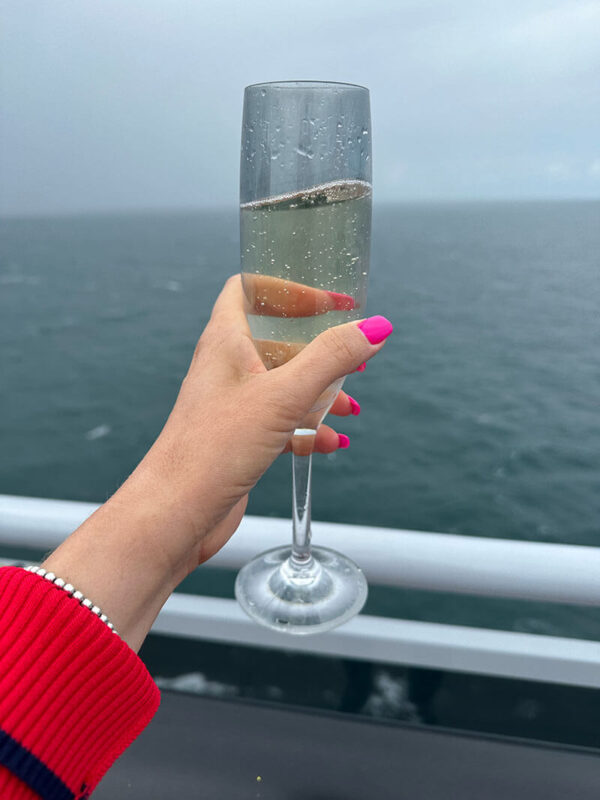 A hand with bright pink nail polish and a beaded bracelet holds up a champagne flute filled with bubbly in front of an open ocean view on a cloudy day.