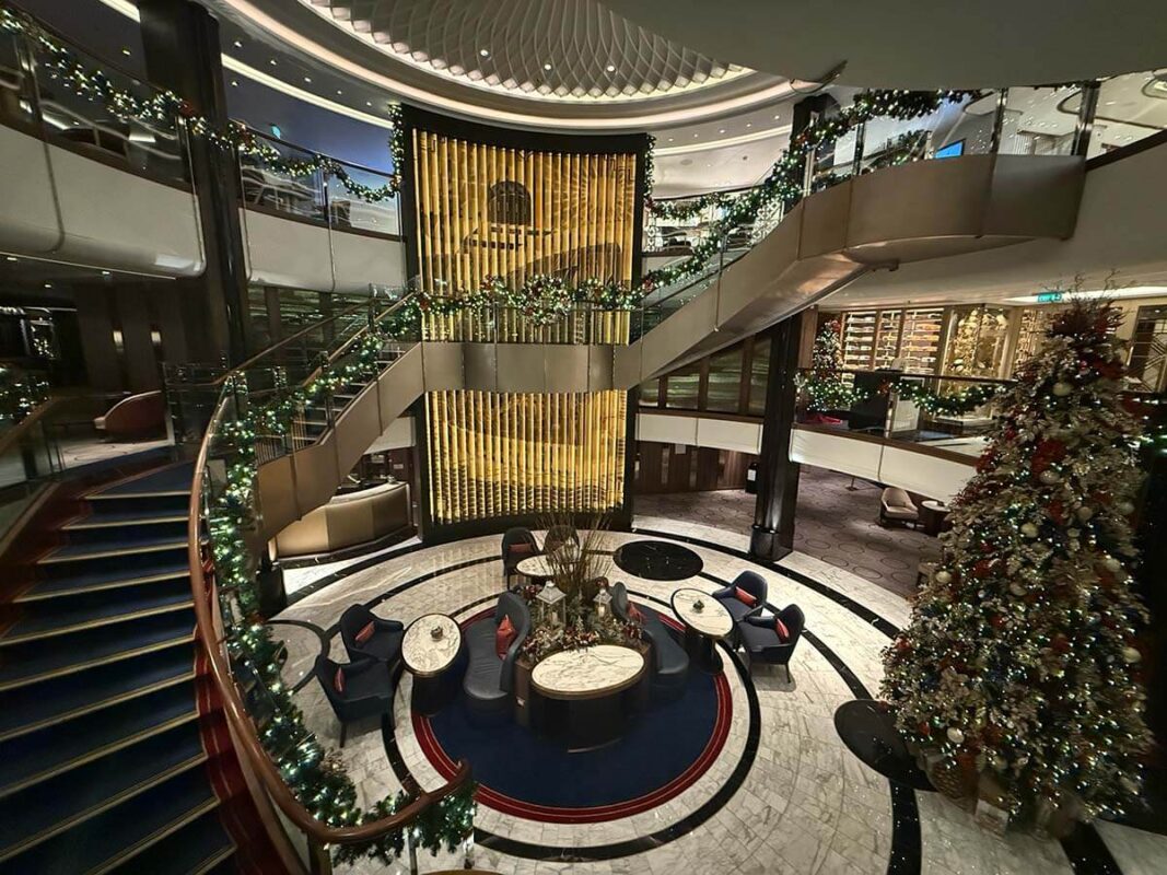 A festive looking atrium on Cunard's Queen Anne featuring a grand staircase, holiday garlands, a towering Christmas tree, and elegant seating beneath a golden vertical art piece.