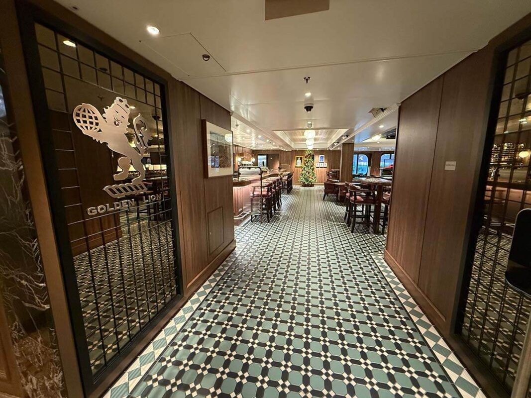 Entrance to the Golden Lion pub on Cunard’s Queen Anne, with a black and white tiled floor, traditional wooden decor, pub-style tables and chairs, and a Christmas tree at the far end.