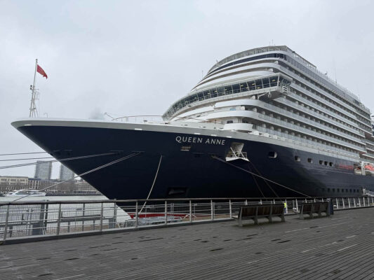 Cunard’s Queen Anne docked in Rotterdam with its navy hull, white balconies, and prominent bow with the ship’s name in bold white letters.