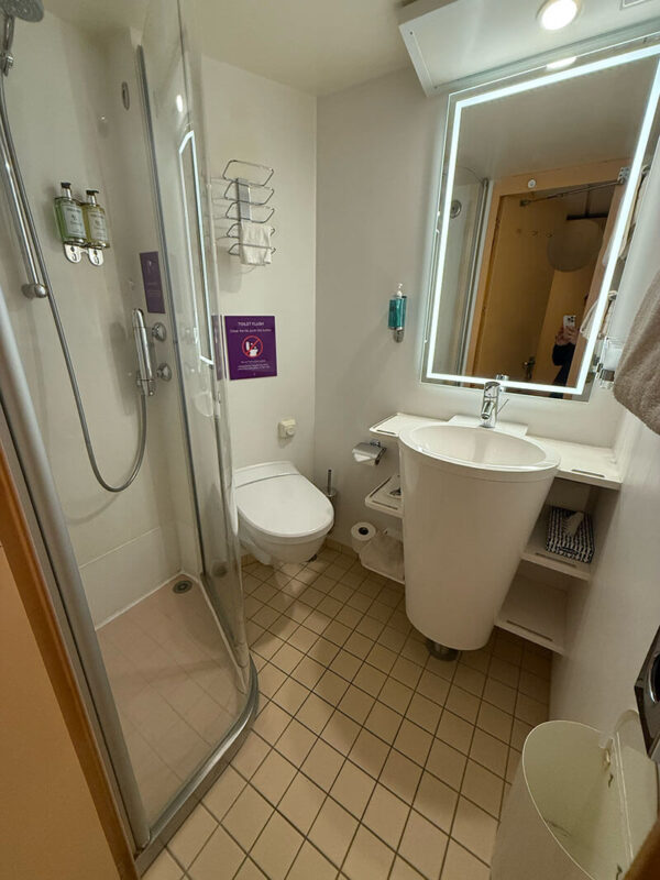 Compact cruise ship bathroom in an inside cabin on Ambassador Ambition with a glass shower, wall mounted toiletries, small sink with illuminated mirror, and a toilet.