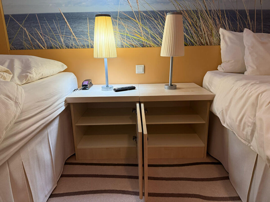 Two single beds in an inside cabin on Ambassador Ambition separated by a bedside table with two lamps, a phone, and a remote, with a wall mural of a beach scene behind them.