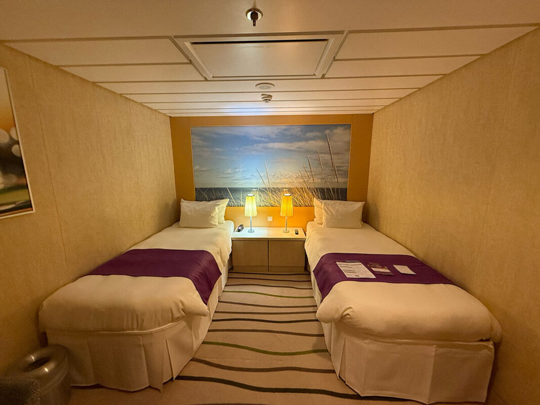 Two single beds in an inside cabin on Ambassador Ambition with purple runners, bedside lamps, and a central cabinet under a beach mural.