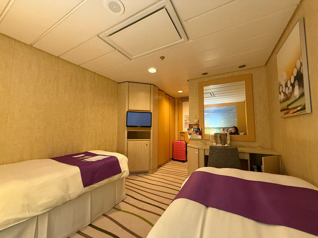 Inside cabin on Ambassador Ambition with two single beds, purple bed runners, a wall mounted TV, desk with mirror, and a pink suitcase near the entrance.