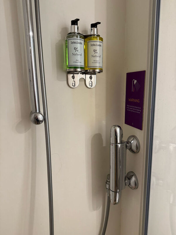 Close up of shower controls and wall mounted shampoo and body wash dispensers in an inside cabin bathroom on Ambassador Ambition with a purple warning sign.