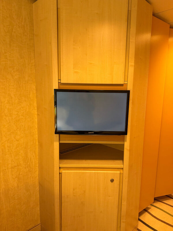 Wall mounted TV in an inside cabin on Ambassador Ambition set into a wooden unit with shelving and a closed cabinet below.