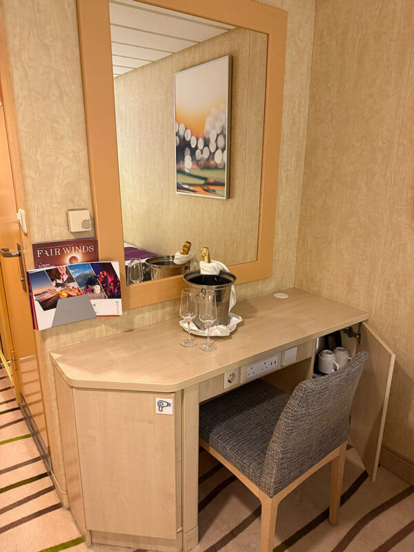 Vanity desk in an inside cabin on Ambassador Ambition with a mirror, chair, champagne bottle in an ice bucket, two glasses, and a brochure titled "FAIR WINDS."