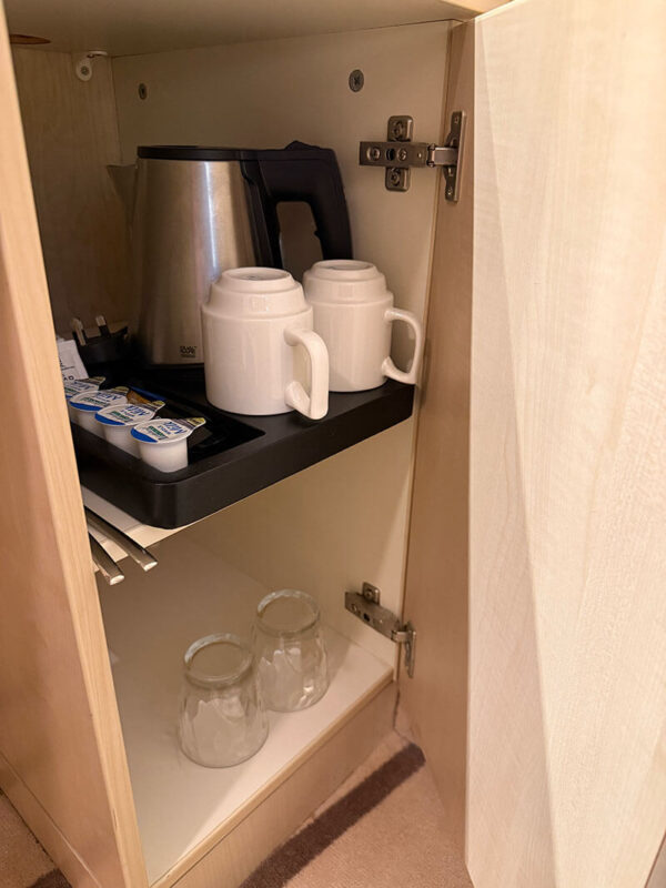 Open cabinet in an inside cabin on Ambassador Ambition with an electric kettle, two white mugs, teaspoons, and packets of coffee and milk, with two drinking glasses stored on the lower shelf.