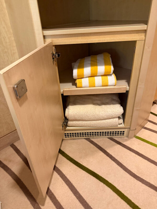 Open storage cabinet in an inside cabin on Ambassador Ambition with folded white towels and yellow striped pool towels neatly stacked on shelves.