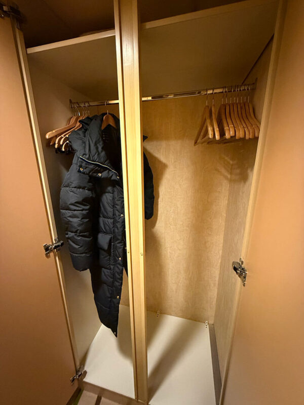 Open wardrobe in an inside cabin on Ambassador Ambition with hanging space, wooden hangers, and a black coat on one side.