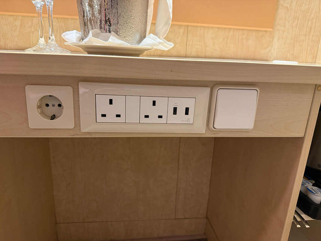Power outlet panel in an inside cabin on Ambassador with European and UK sockets, two USB ports, and a light switch beneath a desk holding glasses and an ice bucket.