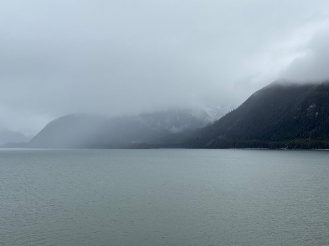 Dark forested mountains rise from the calm grey water along the Alaskan coastline. Thick clouds and mist obscure the mountain peaks during an overcast day on an Alaska cruise.
