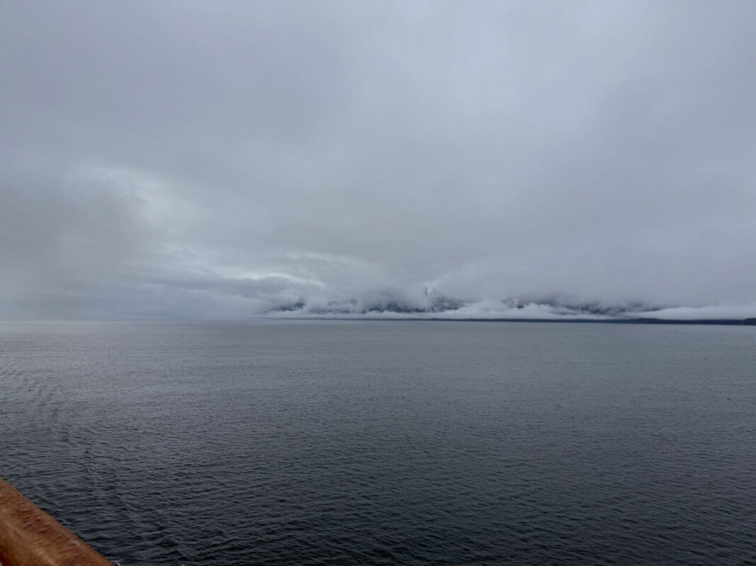 A vast expanse of grey water stretches towards distant mountains almost completely hidden beneath thick low clouds. The gloomy weather creates a very different view of Alaska from the dramatic mountain scenery often seen in travel photographs.