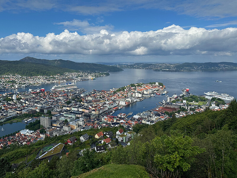 Panoramic view of Bergen in Norway from the top of the Fløibanen funicular railway. Colourful buildings surround the harbour with several large cruise ships docked along the waterfront and green hills stretching into the distance beneath a blue sky filled with white clouds.