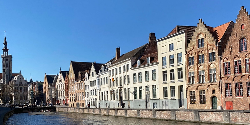 A row of historic brick and whitewashed buildings with stepped gables lines a canal in Bruges Belgium. The medieval architecture reflects the character of the old town with a tall bell tower visible in the distance beneath a clear blue sky.