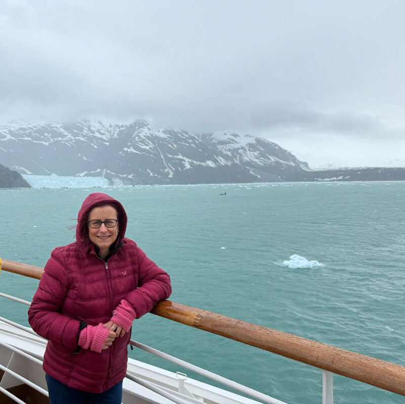 Emmaonline wearing a burgundy hooded jacket and pink gloves leans against the railing of a cruise ship in Alaska. Behind her are turquoise waters dotted with small pieces of ice and a glacier beneath snow covered mountains and low grey clouds.