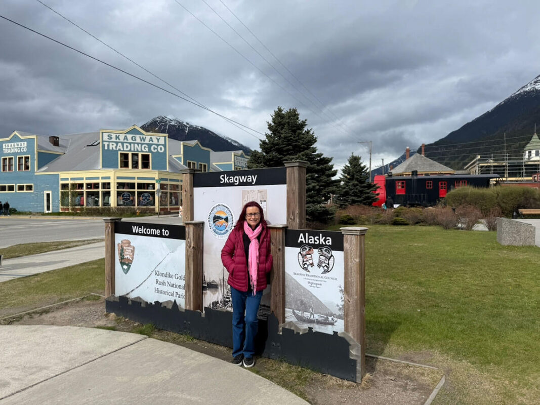 Emmaonline with red hair wearing a burgundy jacket and bright pink scarf stands smiling beside the "Welcome to Skagway Alaska" sign. Colourful historic buildings and snow capped mountains are visible behind her.