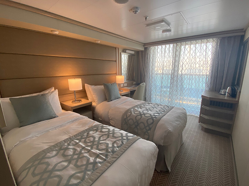 Cruise ship balcony cabin with two single beds dressed in white bedding and grey patterned throws. The room features bedside lamps, a small desk, storage drawers and floor to ceiling glass doors leading to a private balcony.