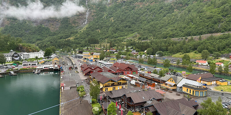 levated view over the waterfront in Flåm Norway with colourful wooden buildings, a railway station and a long pier beside calm green water. Steep forested mountains rise behind the village with low clouds hanging over the hillside.