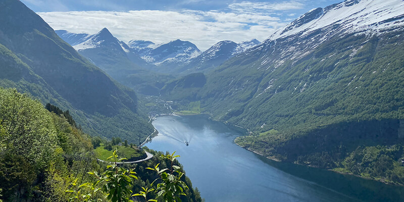Panoramic view of a Geirangerfjord winding between towering green mountains with snow covered peaks. A narrow road curves along the mountainside and a small boat travels through the water far below.