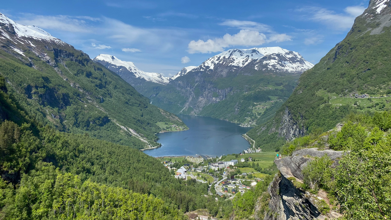 Panoramic view of Geirangerfjord in Norway from a high mountain viewpoint. Deep blue water winds between steep green mountains topped with snow while the village of Geiranger sits in the valley below beneath a bright blue sky.