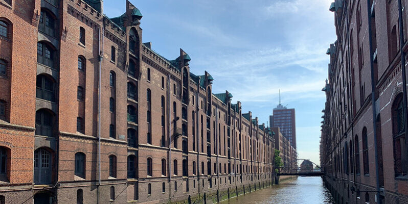 Historic red brick warehouse buildings line both sides of a narrow canal in the Speicherstadt district of Hamburg Germany. A small bridge crosses the water in the distance beneath a blue sky.
