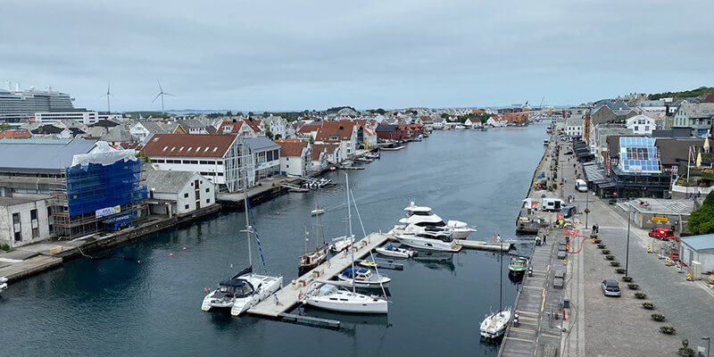 Elevated view over Haugesund harbour in Norway with sailboats and luxury yachts moored along the waterfront. Colourful wooden buildings with red tiled roofs line one side of the water while a road and promenade run along the other beneath an overcast sky.