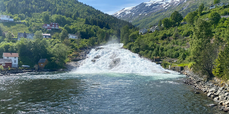 A powerful waterfall rushes down a rocky hillside into a river surrounded by lush green trees and colourful houses in Hellesylt, Norway. Snow covered mountains rise in the background beneath a bright blue sky.