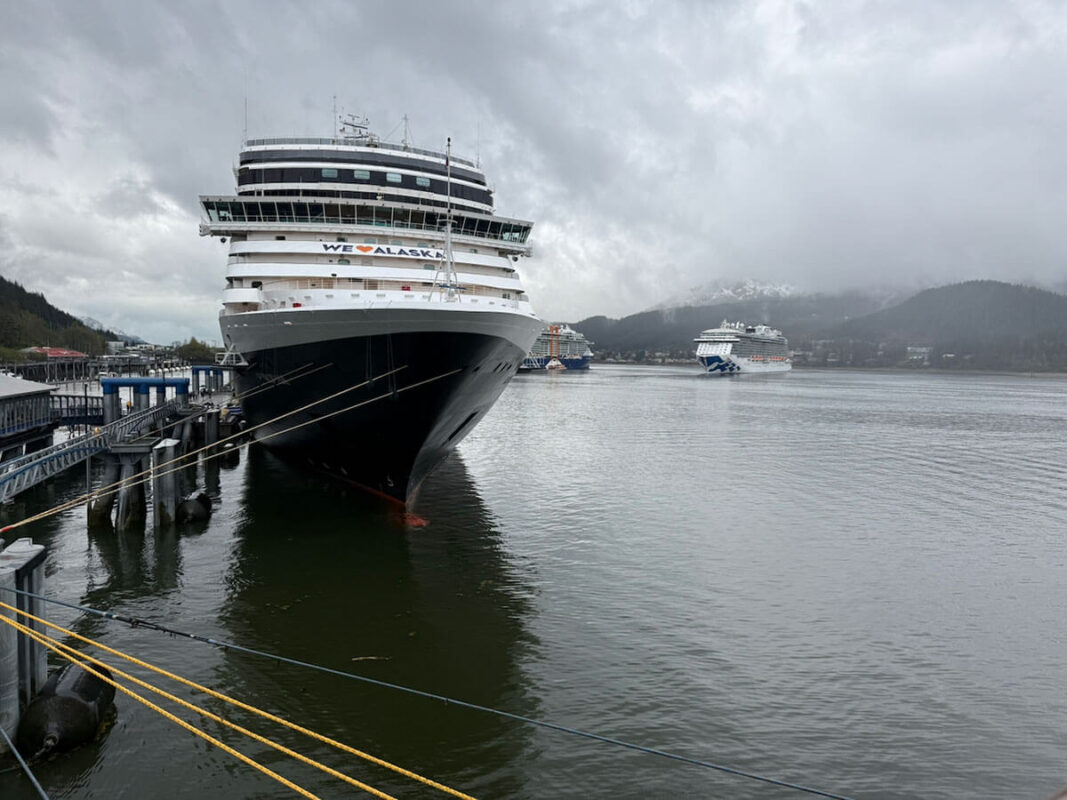 Holland America Koningsdam cruise ship with the words "We Love Alaska" on the front is docked at a port beneath grey cloudy skies. Two other cruise ships are anchored in the distance with misty mountains behind them.