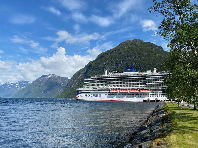 A large white P&O Cruises ship is docked beside a tree lined shoreline in a Norwegian fjord. Towering green mountains with snow covered peaks rise behind the ship beneath a bright blue sky dotted with fluffy white clouds.